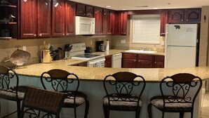 Una cocina con armarios de madera oscura, un refrigerador blanco, una estufa y un lavamanos. Hay tres taburetes de bar con asientos acolchados.