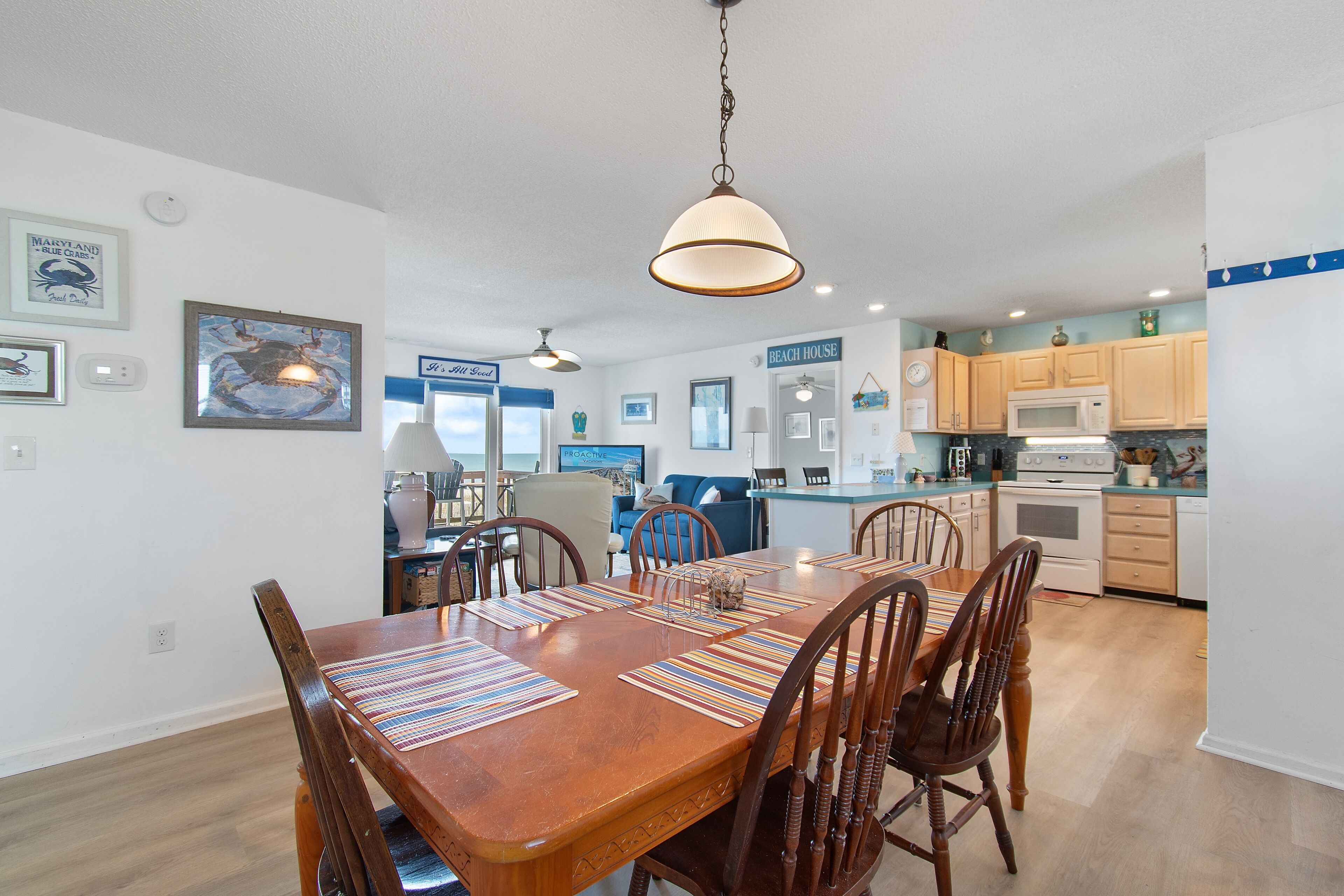 A spacious dining area with a wooden table and chairs, a hanging light fixture, and a kitchen in the background.