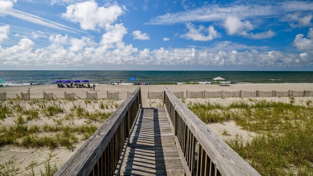 A wooden boardwalk leading to a sandy beach with umbrellas and a few people.