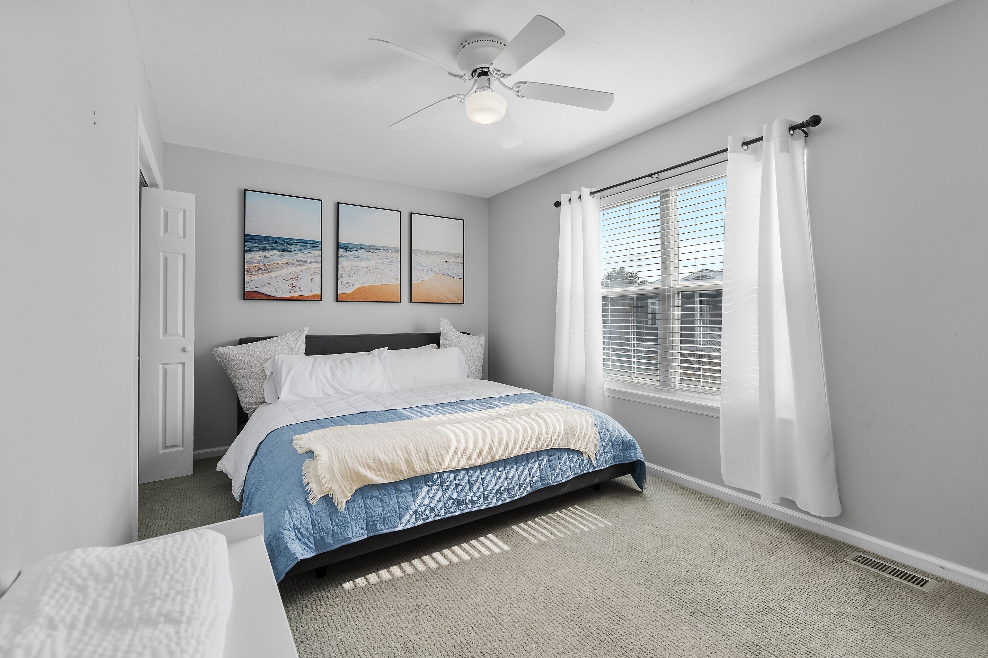 Une chambre à coucher comprenant un lit, un ventilateur de plafond, une fenêtre avec des rideaux et deux œuvres d’art murales sur le thème de la plage.
