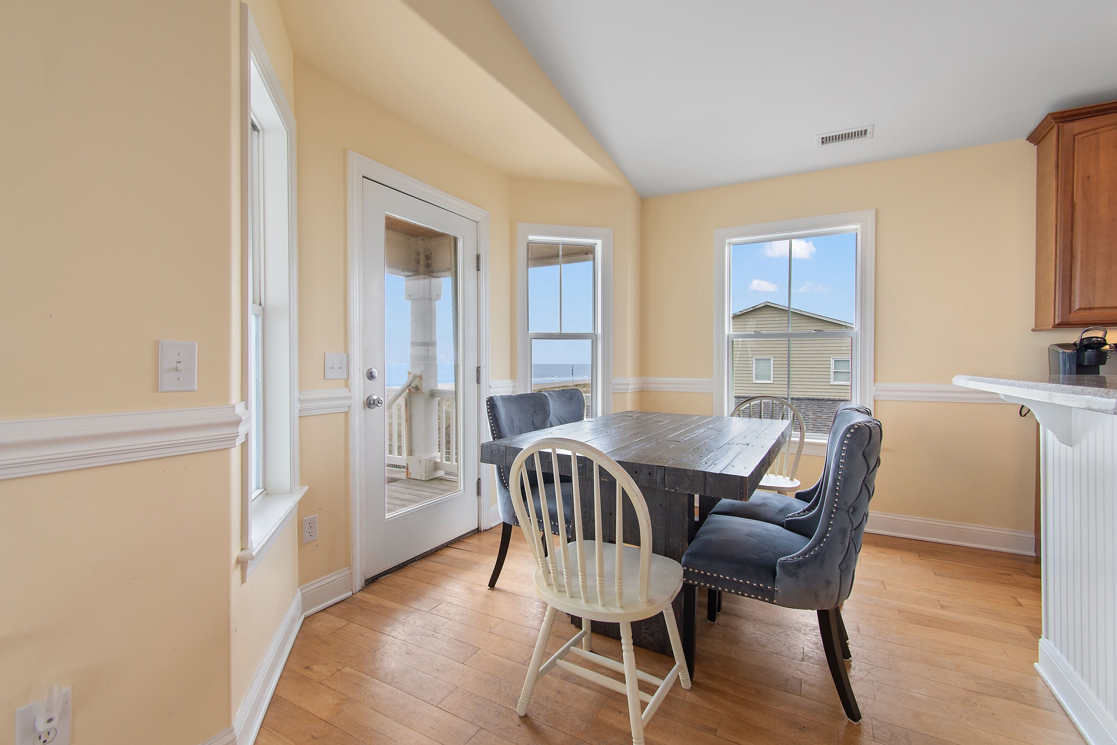 A dining area with a wooden table and chairs, a view through a door, and a kitchen area with cabinets.