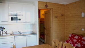 Eine kompakte Küche mit modernen Geräten, ein Essbereich mit Holztisch und -stühlen sowie eine gemütliche Schlafgelegenheit mit einer rot gemusterten Couch.