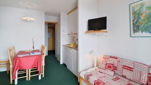 A compact room with a dining table, chairs, a sofa, a television, and a kitchen area.