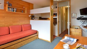 Una habitación acogedora con un sofá rojo estampado, una estantería de madera, una mesa de comedor con una revista y un televisor en la pared.