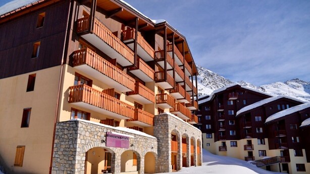 Un hotel de varios pisos con balcones de piedra y madera, una entrada de piedra y una montaña nevada al fondo.