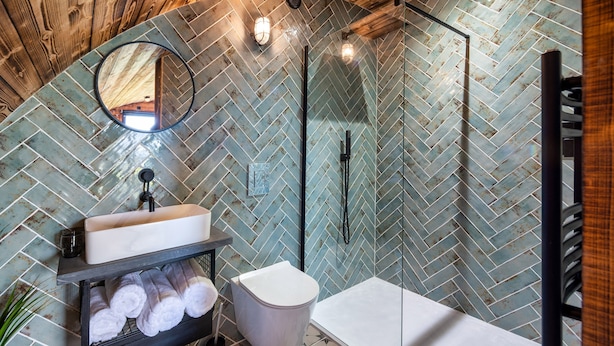 Un baño con patrón de mosaico de espiga, un espejo redondo, un lavamanos blanco, un inodoro y una zona de ducha.