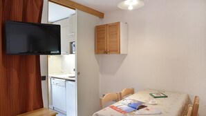 A small room with a bed, a table with a floral tablecloth, a wooden cabinet, a television, and a door leading to a kitchen. - travelski home classic - Résidence 3000 - Studio 4 personnes 529 (La Plagne)