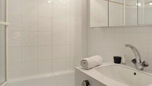 A modern bathroom with white tiles, a white bathtub, a sink with a faucet, and a mirror above the sink. - travelski home classic - Résidence 3000 - Studio 4 personnes 529 (La Plagne)
