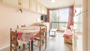 Een eethoek met een tafel voor vier personen, houten stoelen, een bijzettafeltje en een bank. Er is een raam met gordijnen, een televisie op een dressoir en een koelkast.