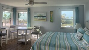 Ein Schlafzimmer mit einem Bett, einem Schreibtisch, Stühlen, einem Deckenventilator und zwei mit Vorhängen versehenen Fenstern.