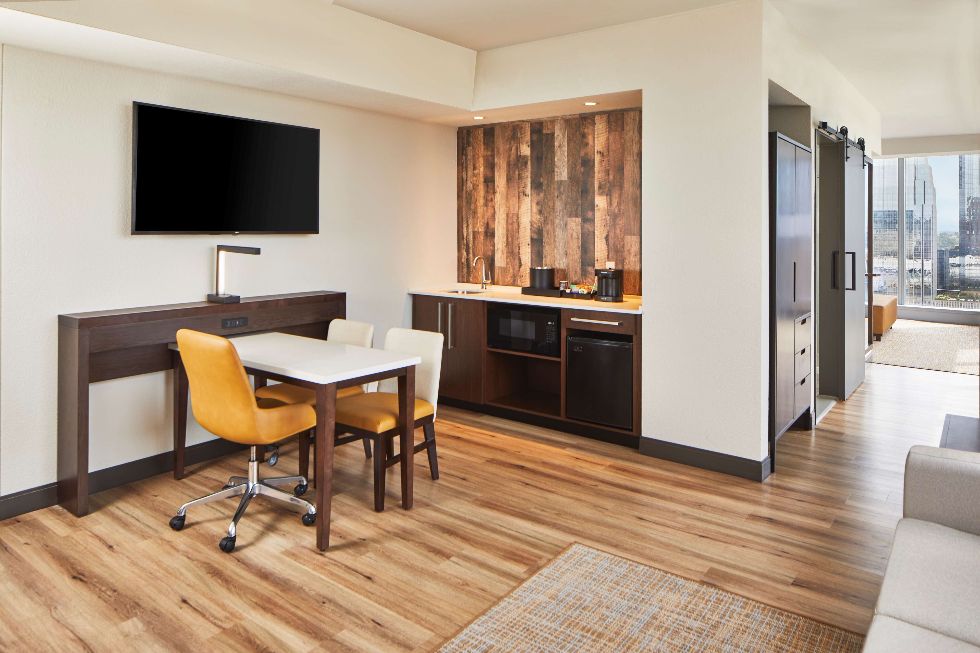A modern hotel room with a dining area, kitchenette, and a large window offering a city view.
