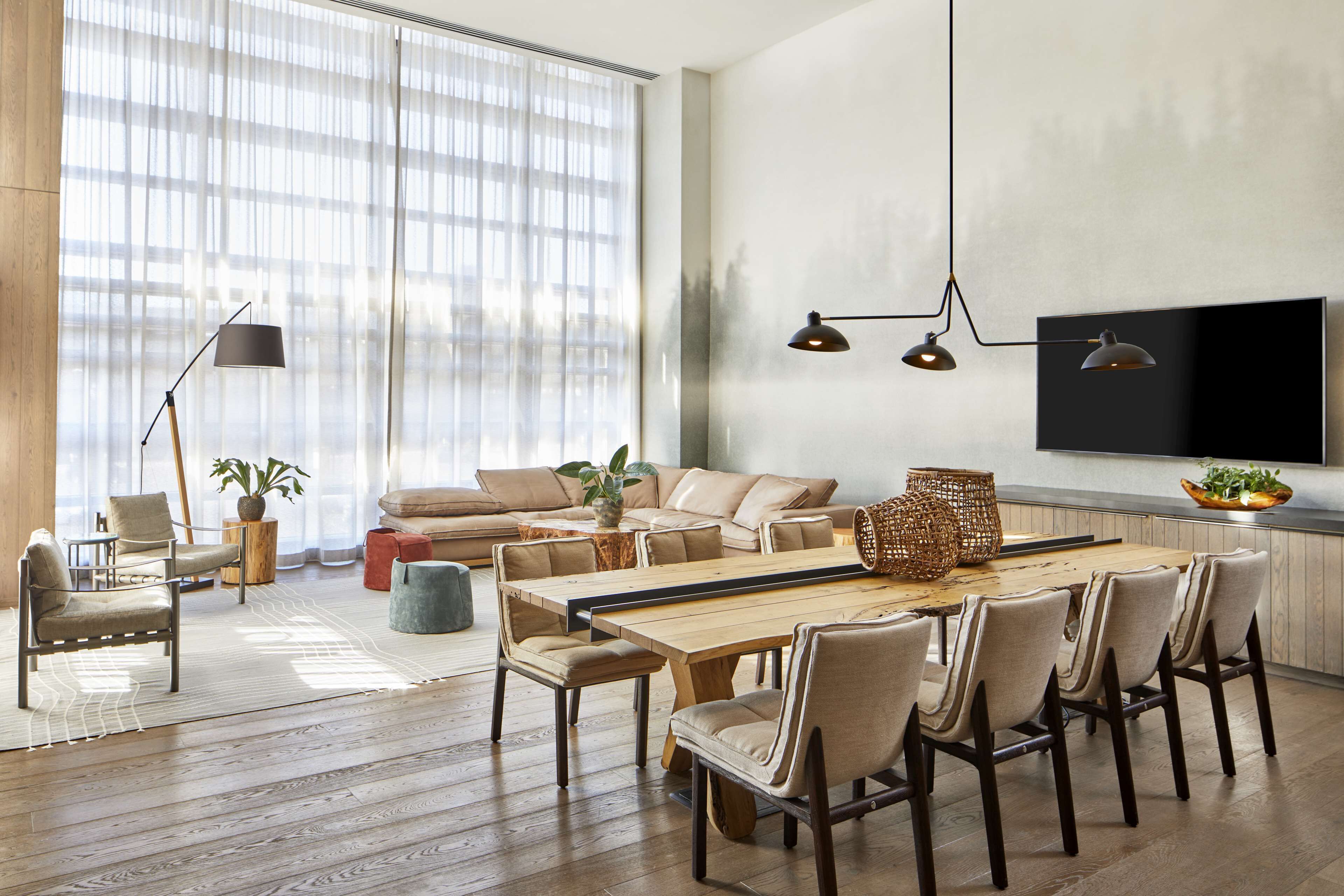 A spacious living room with a large wooden dining table, beige sofas, and a TV mounted on the wall.