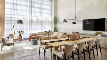 A spacious living room with a large wooden dining table, beige sofas, and a TV mounted on the wall.
