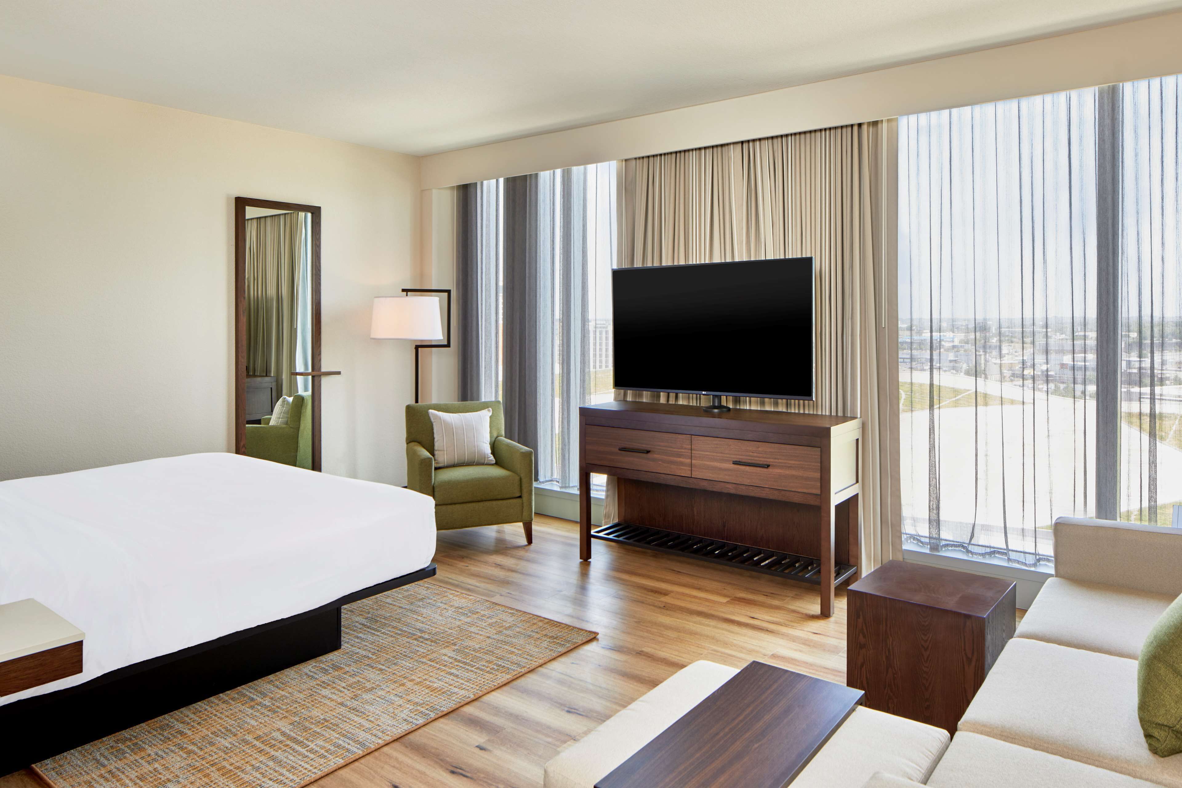 Habitación de hotel con una cama grande, un televisor de pantalla plana, un sofá, una silla y vistas de la ciudad a través de amplias ventanas.