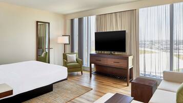 Habitación de hotel con una cama grande, un televisor de pantalla plana, un sofá, una silla y vistas de la ciudad a través de amplias ventanas.
