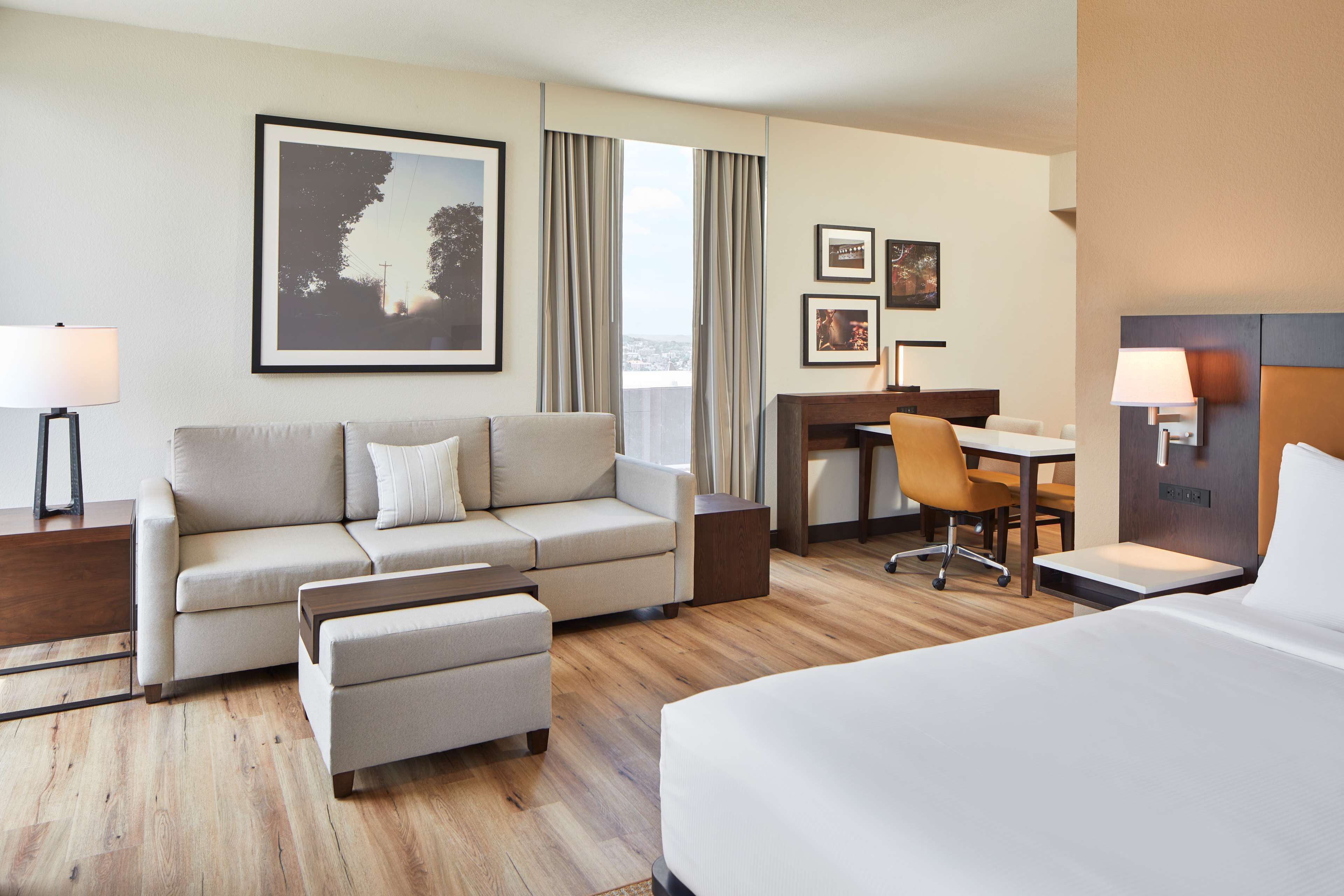 Ein Hotelzimmer mit Bett, Sofa, Schreibtisch und Stuhl. Es gibt ein Fenster mit Vorhängen, ein gerahmtes Bild an der Wand und eine Lampe auf dem Nachttisch.