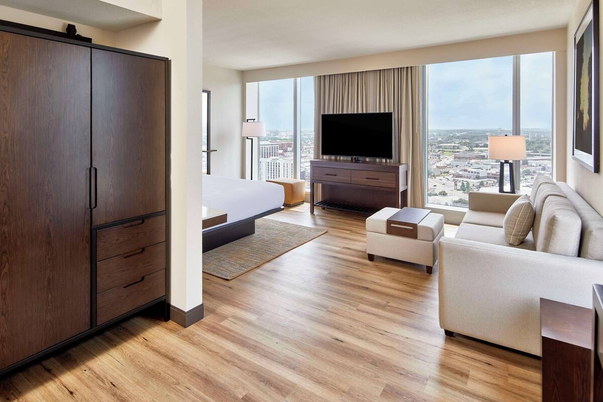A hotel room with a large wardrobe, a bed, a TV, a sofa, and a view of the city.
