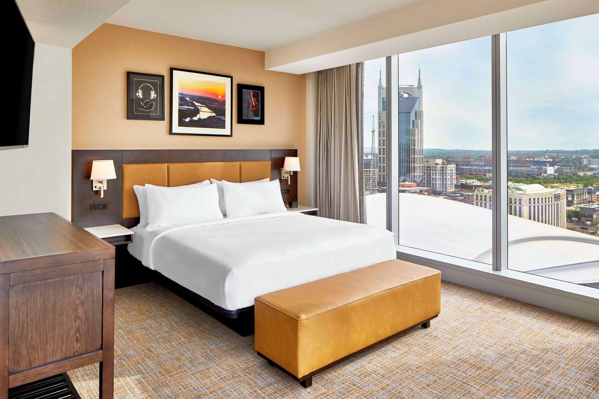 A hotel room with a large bed, a wooden nightstand, a bench, and a city view through large windows.