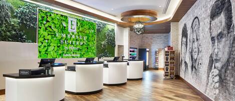 A hotel reception area with a green wall displaying the hotel's name and logo, multiple reception desks, a brick wall mural, and a wooden floor.