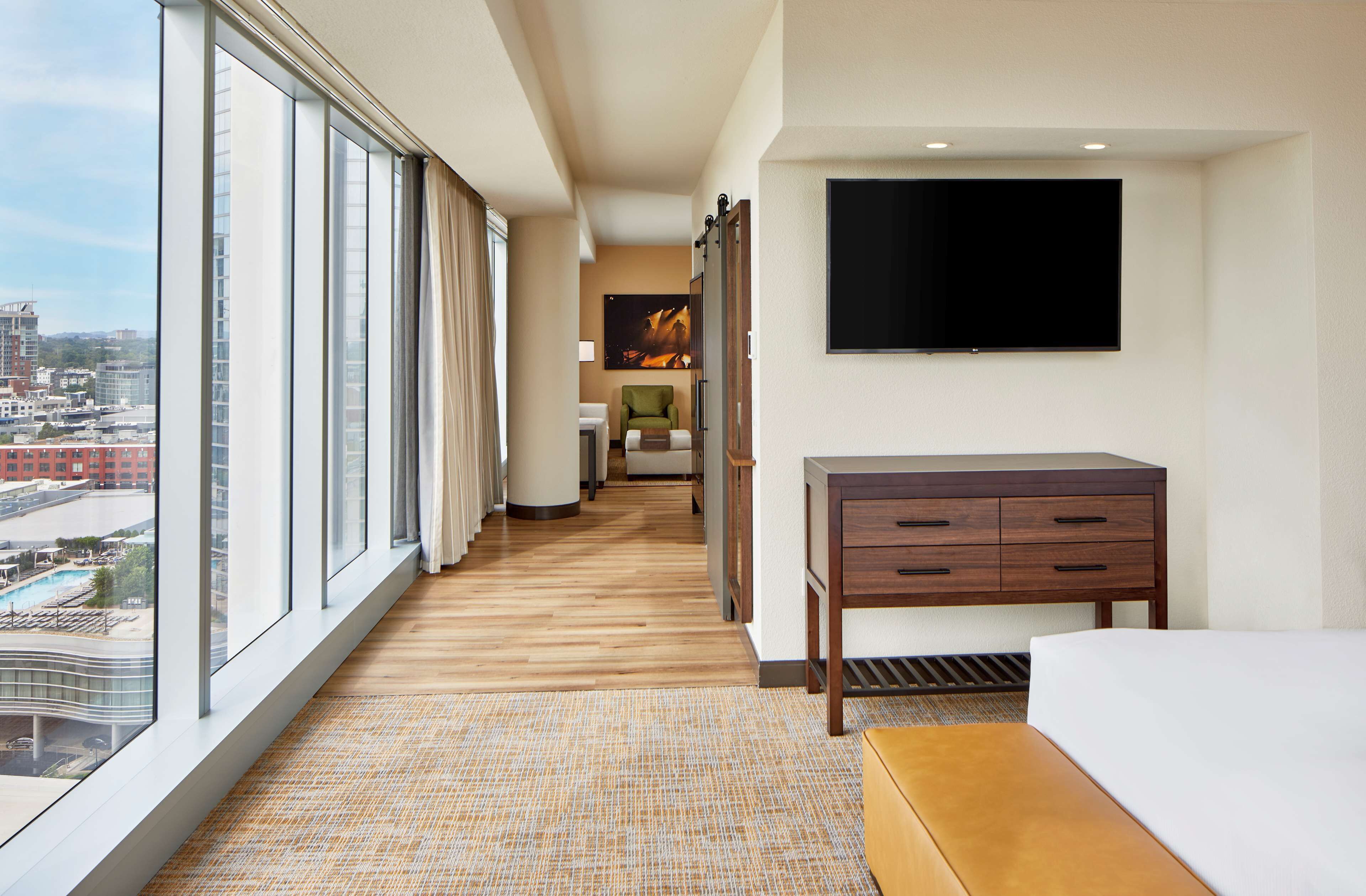 A hotel room with a large window, a bed, a desk, a chair, and a TV.
