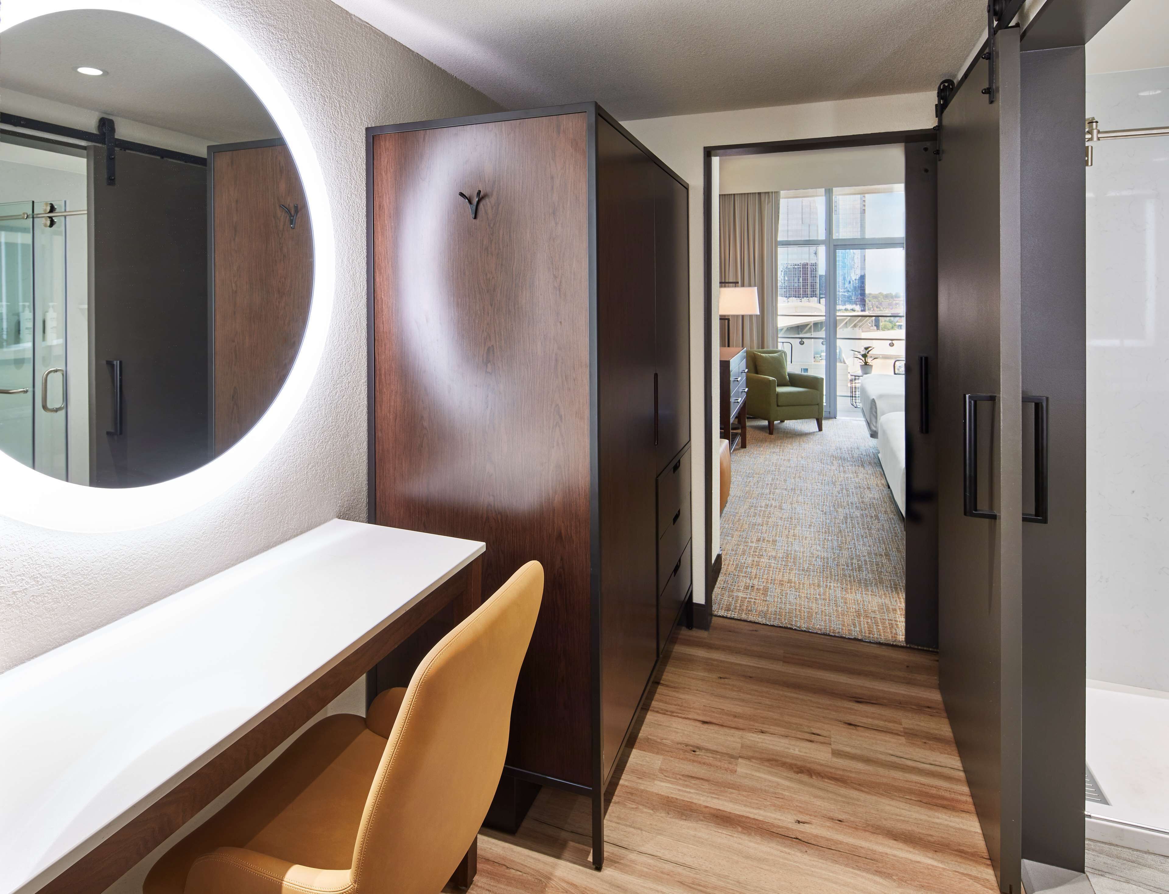Habitación de hotel con un escritorio de madera, un espejo redondo, una silla verde y vistas a la ciudad a través de amplios ventanales.