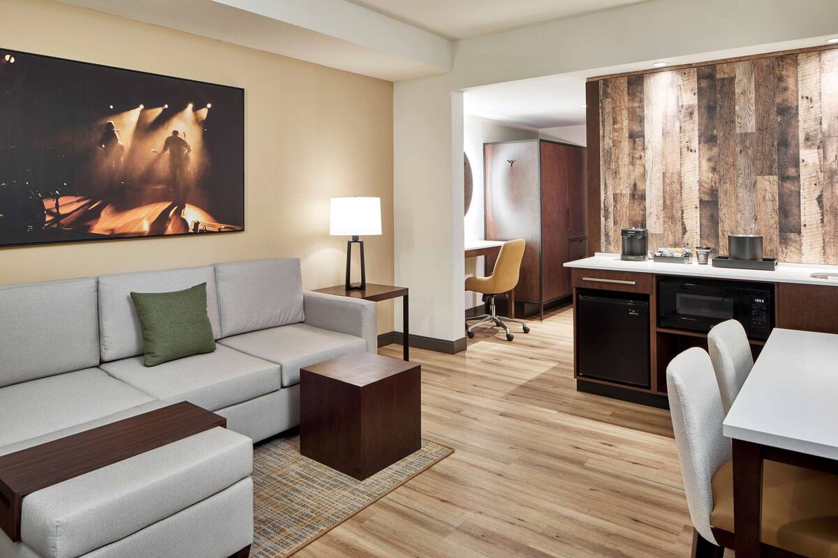 A modern hotel room with a sofa, a coffee table, a lamp, a dining table with chairs, a kitchen area with appliances, and a large painting on the wall.
