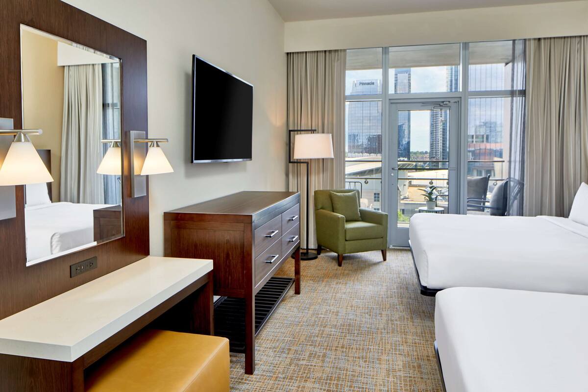 A hotel room with a large bed, a desk with a mirror, a television, and a view of the city through large windows.