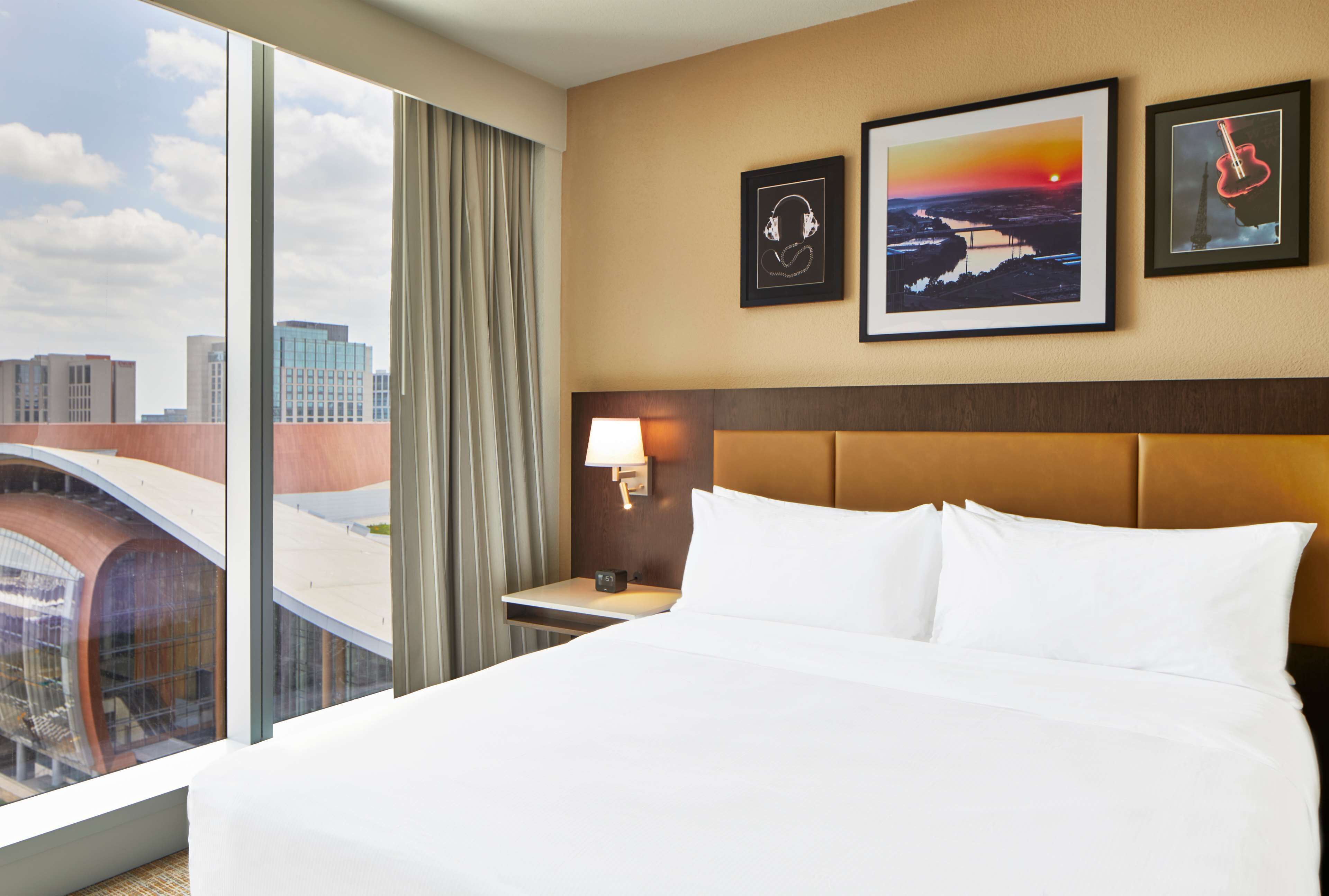 Una habitación de hotel con una cama grande, una mesita de noche con una lámpara y vistas a un paisaje urbano a través de ventanas amplias.