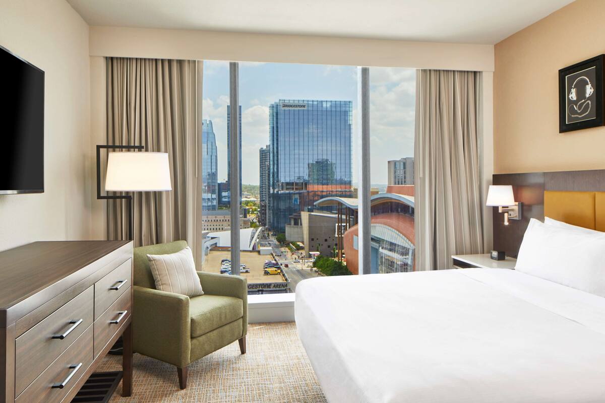 A hotel room with a large bed, a green armchair, a TV, and a view of a cityscape.
