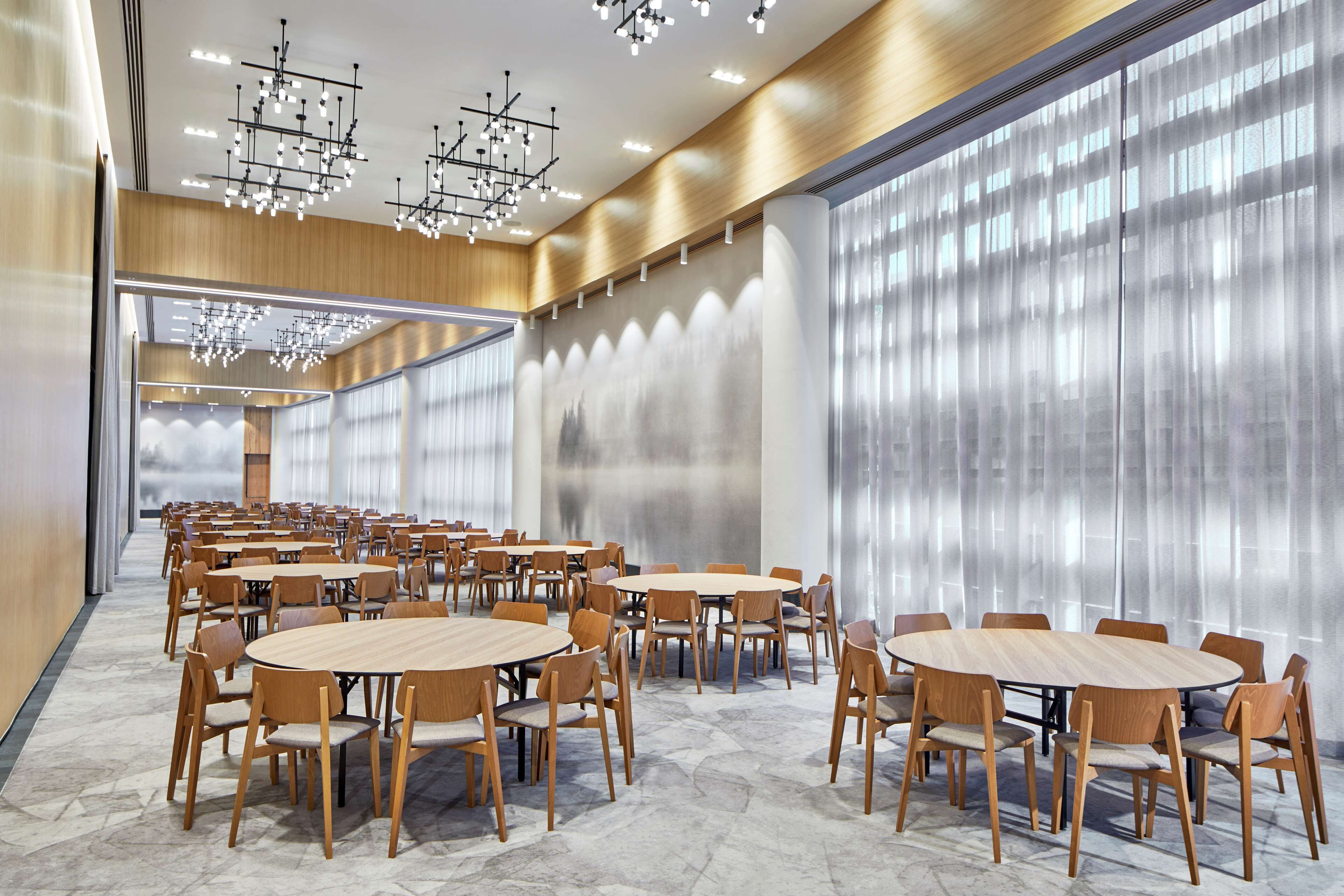 Una amplia sala de conferencias con varias mesas redondas y sillas, ventanas grandes con cortinas translúcidas e iluminación moderna.