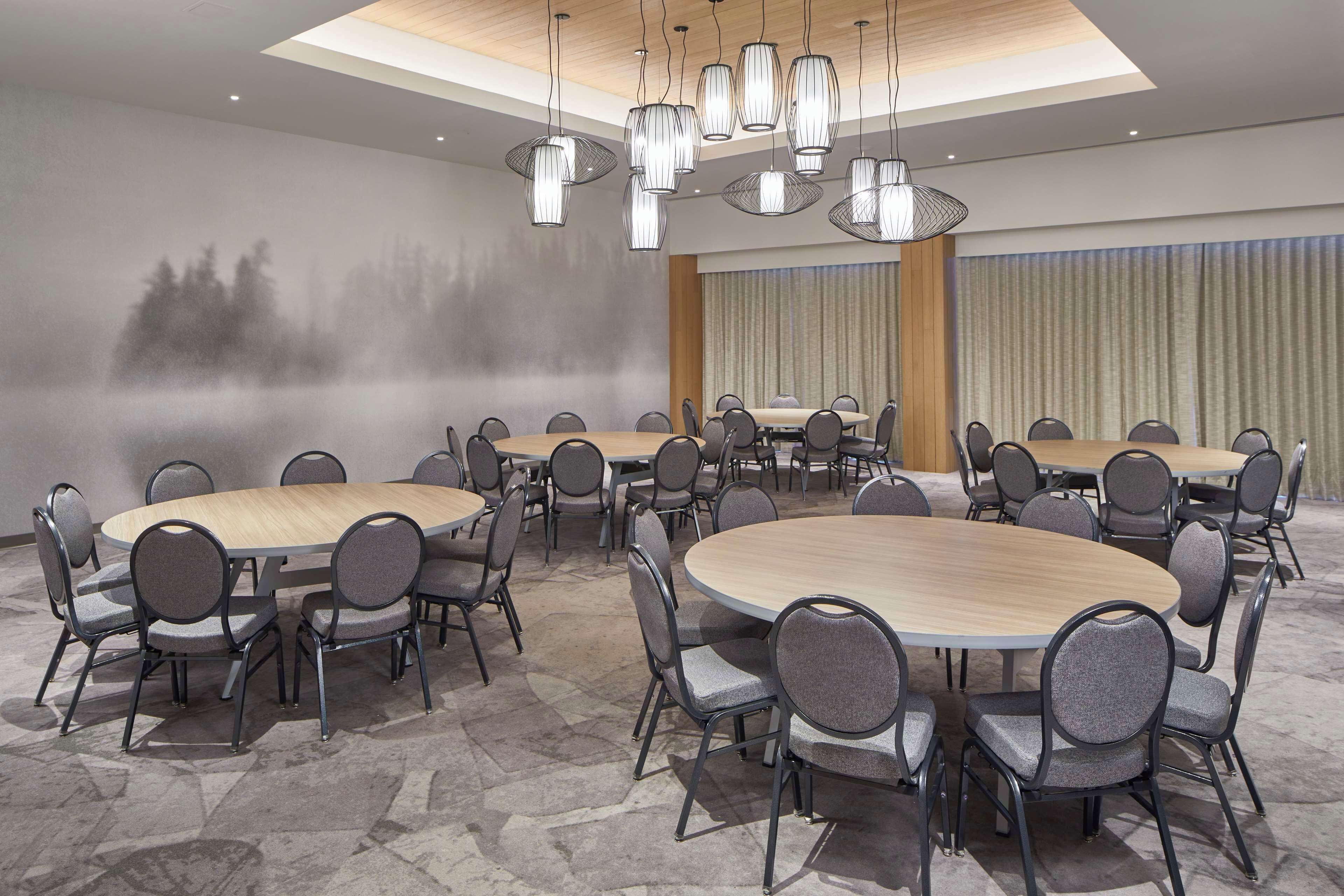 Une salle de conférence dotée de tables et de chaises rondes, d’un grand lustre et d’un mur orné d’une fresque représentant une forêt.
