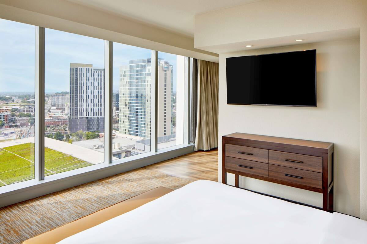 A hotel room with a large window, a bed, a wooden dresser, and a flat-screen TV mounted on the wall.