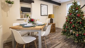 Un coin repas avec une table en bois dressée pour deux personnes, des chaises blanches, un sapin de Noël et une carte accrochée au mur.