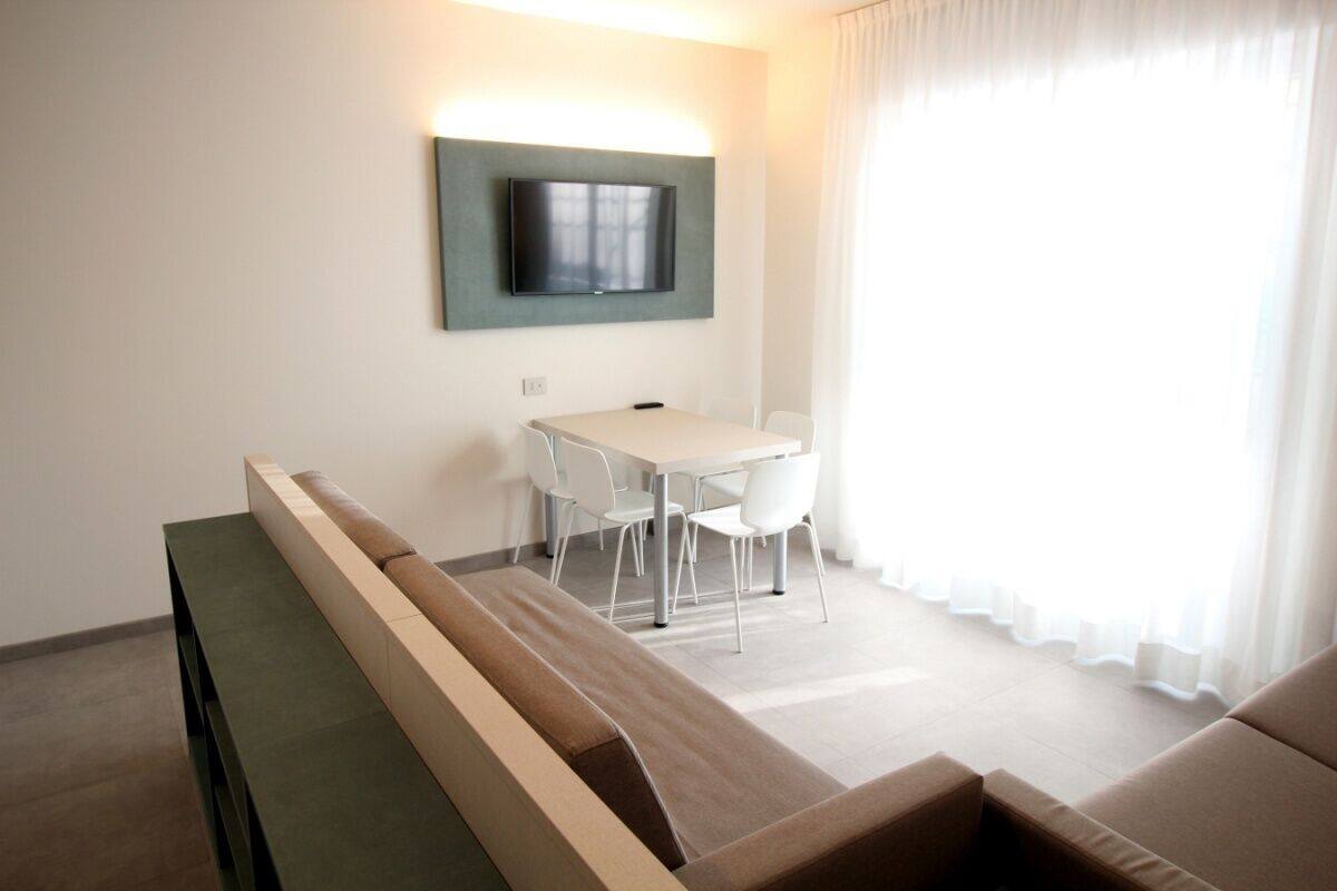 Une chambre d’hôtel moderne équipée d’un canapé, d’une table à manger avec des chaises, d’un téléviseur à écran plat fixé au mur et d’une grande fenêtre avec des rideaux.