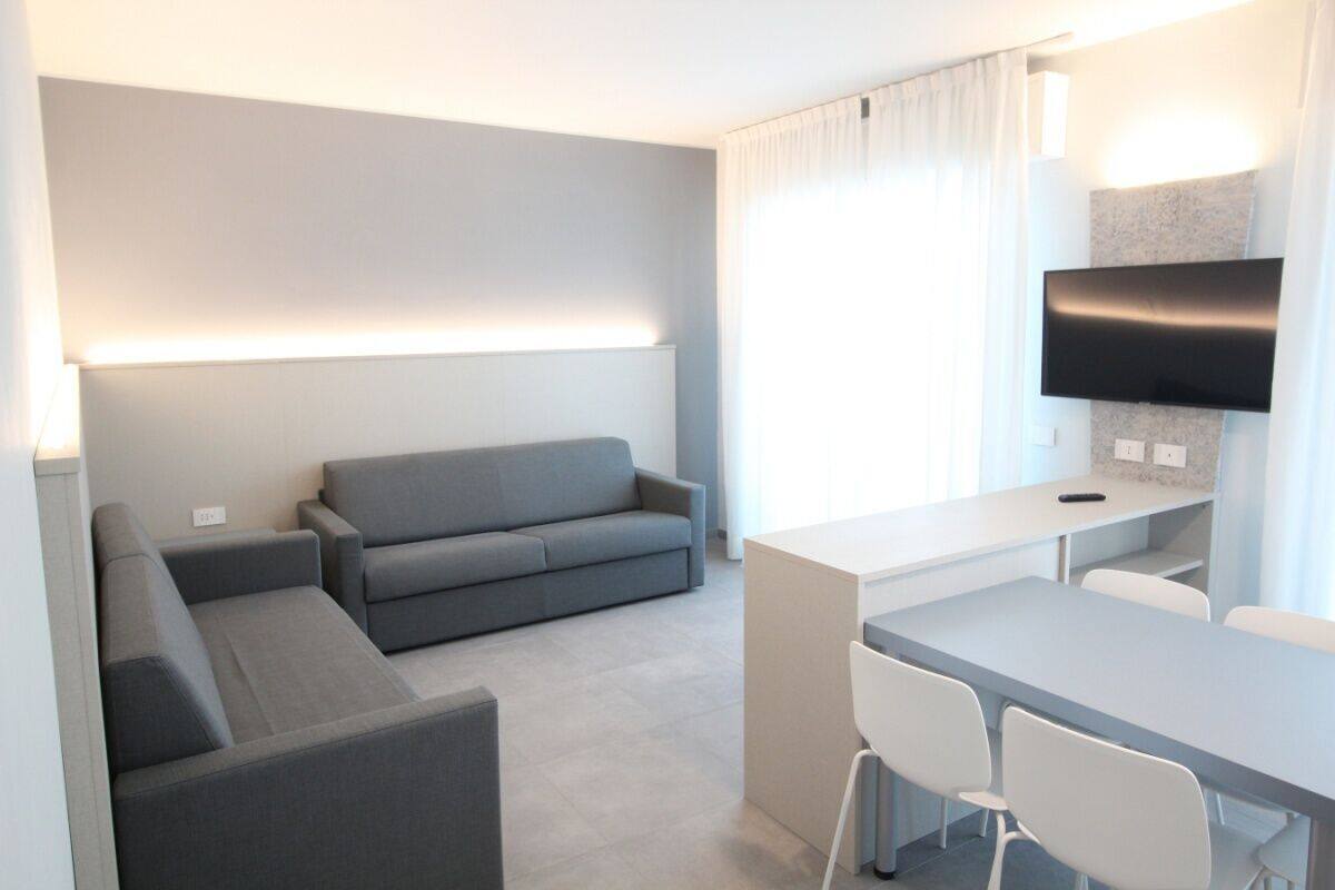 Une chambre d’hôtel moderne avec un canapé gris, une petite table à manger avec des chaises, un téléviseur à écran plat fixé au mur et une grande fenêtre avec des rideaux.