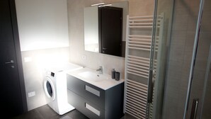A modern bathroom with a built-in washing machine, a large mirror, and a glass shower enclosure.