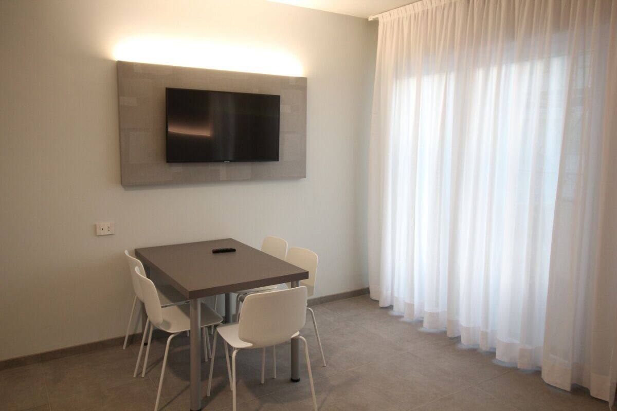 Une chambre moderne dotée d’un téléviseur à écran plat fixé au mur, d’une table à manger avec des chaises et d’une grande fenêtre ornée de rideaux légers.