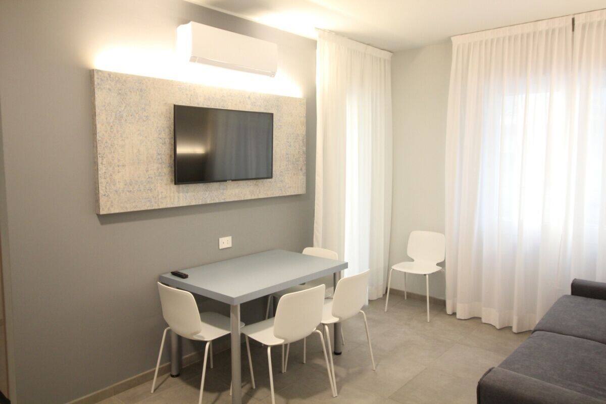 Un salon moderne avec un canapé gris, une table à manger entourée de quatre chaises blanches, un téléviseur à écran plat fixé à un mur texturé et une grande fenêtre ornée de rideaux légers.