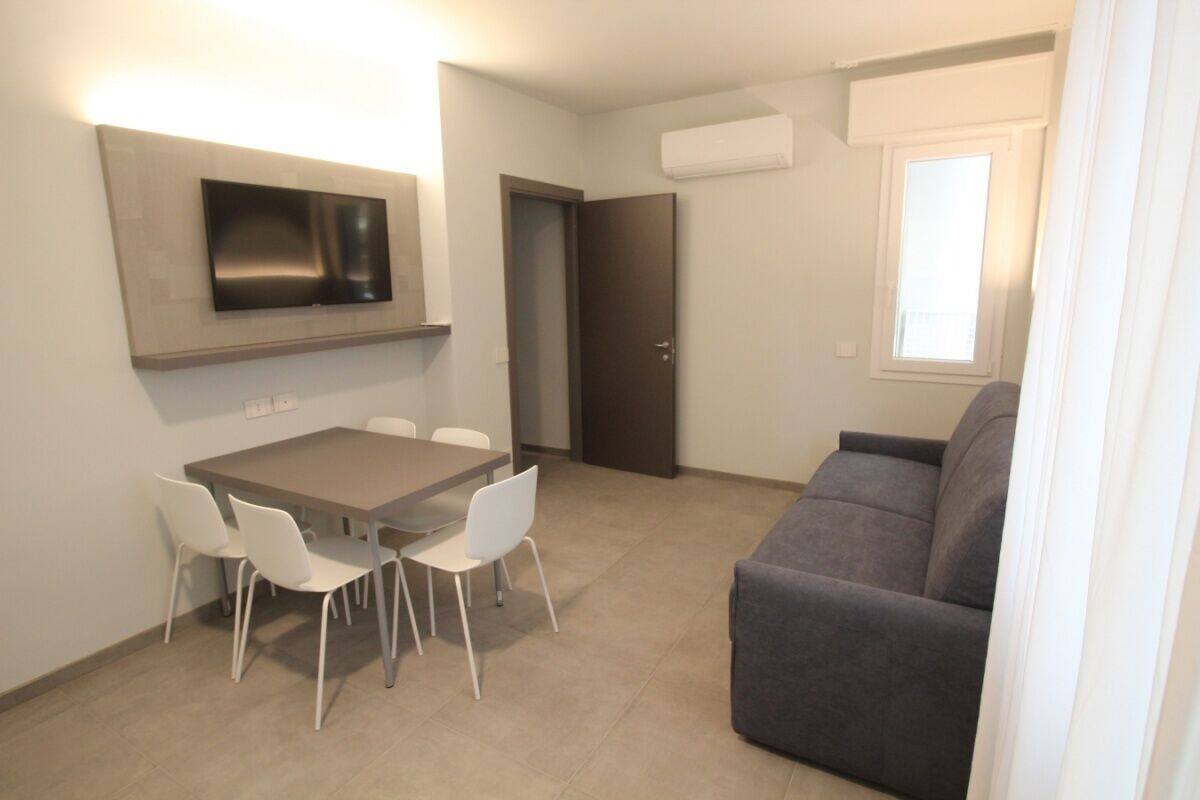Un salon moderne avec un canapé gris, une table à manger avec quatre chaises, un téléviseur à écran plat fixé au-dessus de la table et une fenêtre avec des stores.
