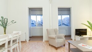 A modern living room with a sofa, a dining table, and a TV. There are two windows with city views. - Apartment SF C - 3 bedrooms 5 pax - 300 meters from Sagrada Familia - FREE WIFI (Barcelona)