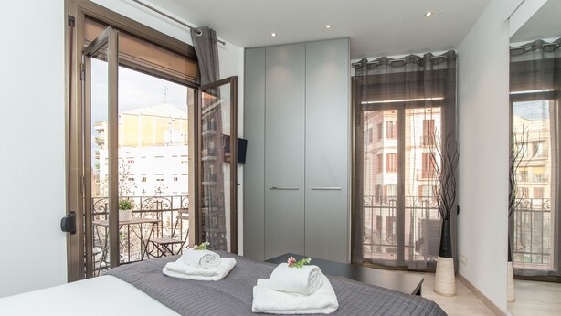 A hotel room with a bed, a TV, a balcony with a table and chairs, and a view of buildings. - Gaudí Views- Suite with view on Hospital Sant Pau (UNESCO Heritage)  - FREE WIFI (Barcelona)