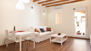 Un salon moderne avec un canapé blanc, une table à manger et une table basse. Le plafond est en bois et une porte donne accès à un espace extérieur agrémenté de plantes en pot.