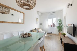 A modern dining area with a glass table, white chairs, a sofa with cushions, a small round table, and a television on a wooden stand.