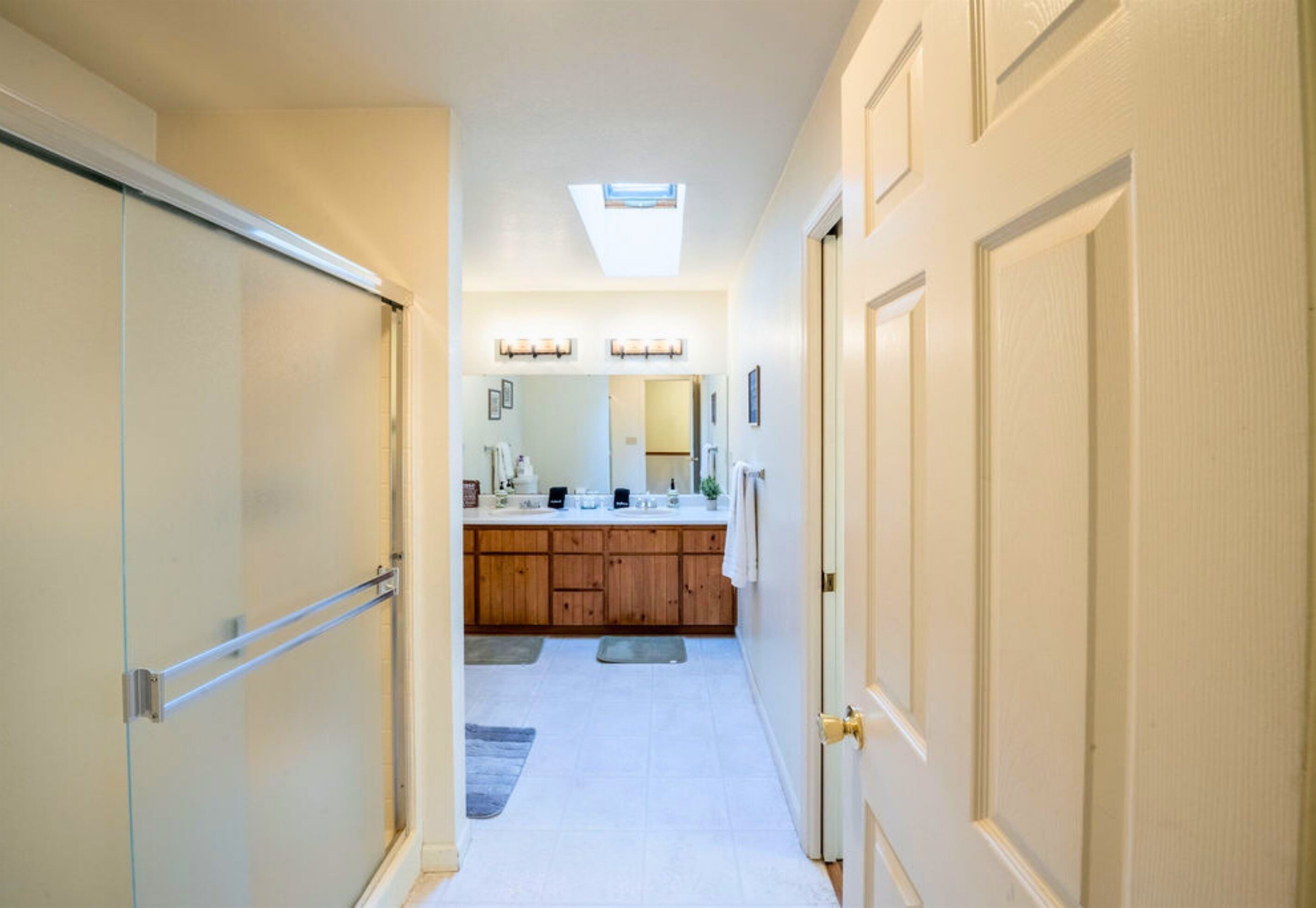 A narrow hallway leading to a bathroom with a skylight, wooden cabinets, and a large mirror.