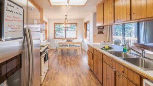 A kitchen with wooden cabinets, a stainless steel refrigerator, a dining table with chairs, and a window with a view of trees. - Serenity in the Pines: Cozy Mountain Retreat (Twain Harte)