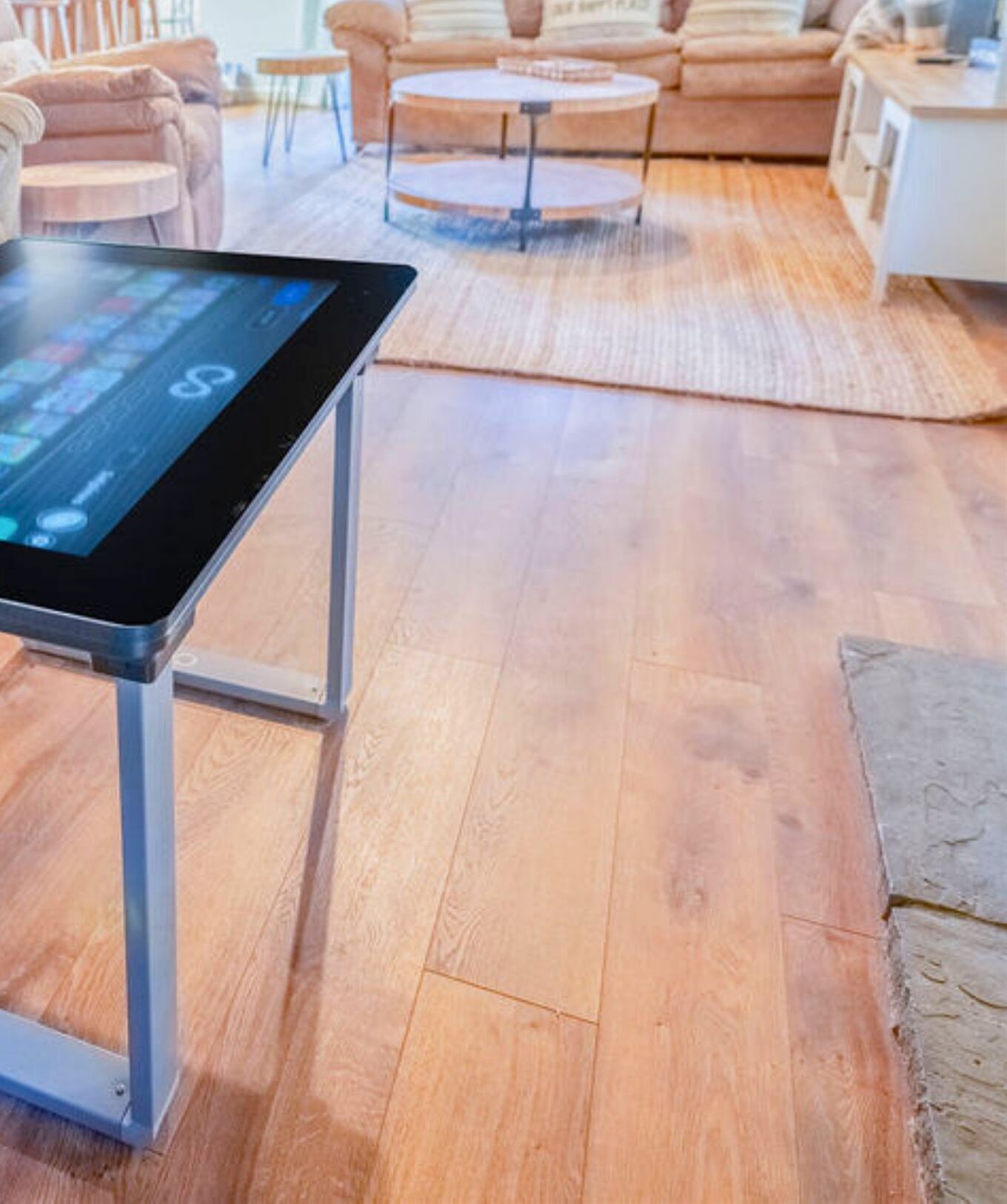 Um tablet em um suporte em sala de estar com piso de madeira, uma mesa de centro redonda e um sofá ao fundo.