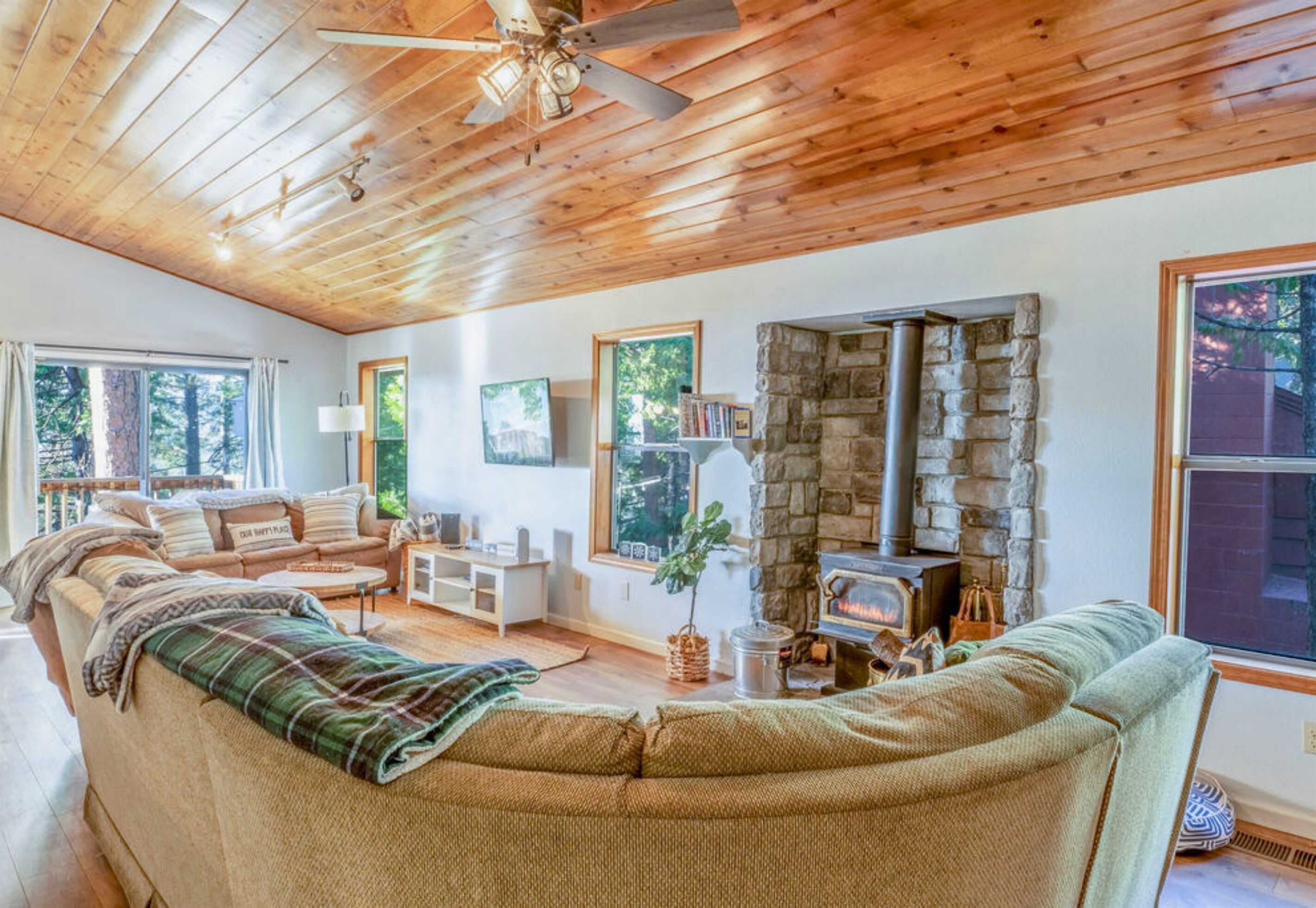 A cozy living room with a stone fireplace, wooden ceiling, and a large window overlooking a forest.