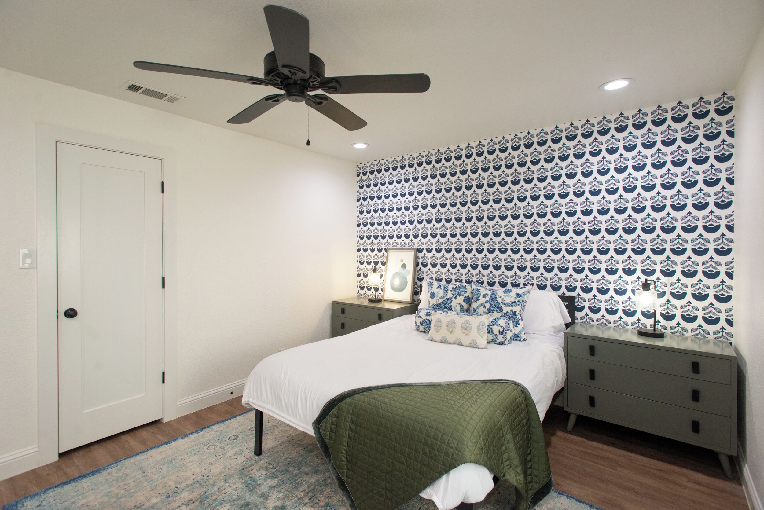 A bedroom with a bed, a nightstand, a ceiling fan, and patterned wallpaper.