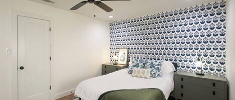 A bedroom with a bed, a nightstand, a ceiling fan, and patterned wallpaper.