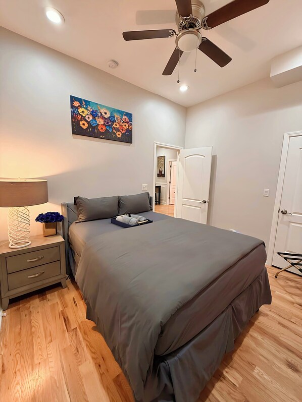 Un dormitorio con cama, mesita de noche, lámpara, ventilador de techo y un cuadro en la pared.